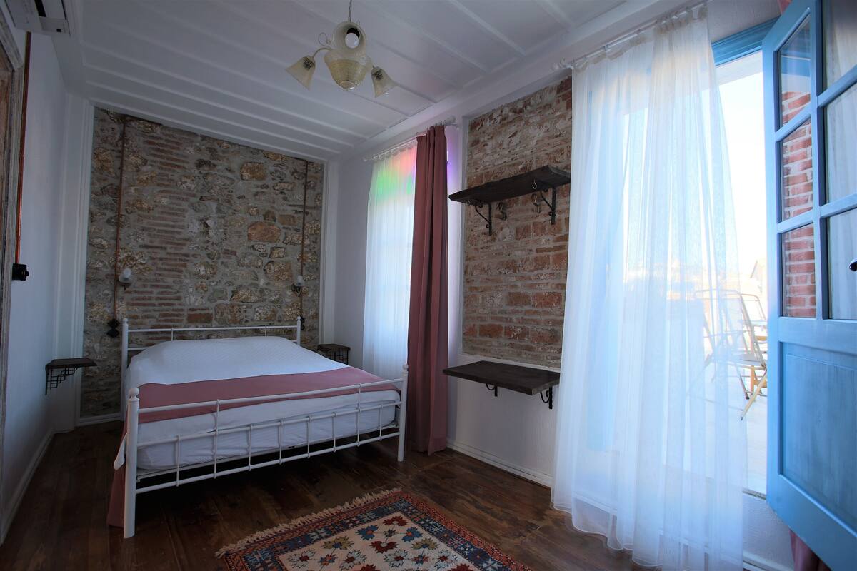 A bedroom with a brick wall, a bed with a white frame, a wooden floor, and a window with sheer curtains.