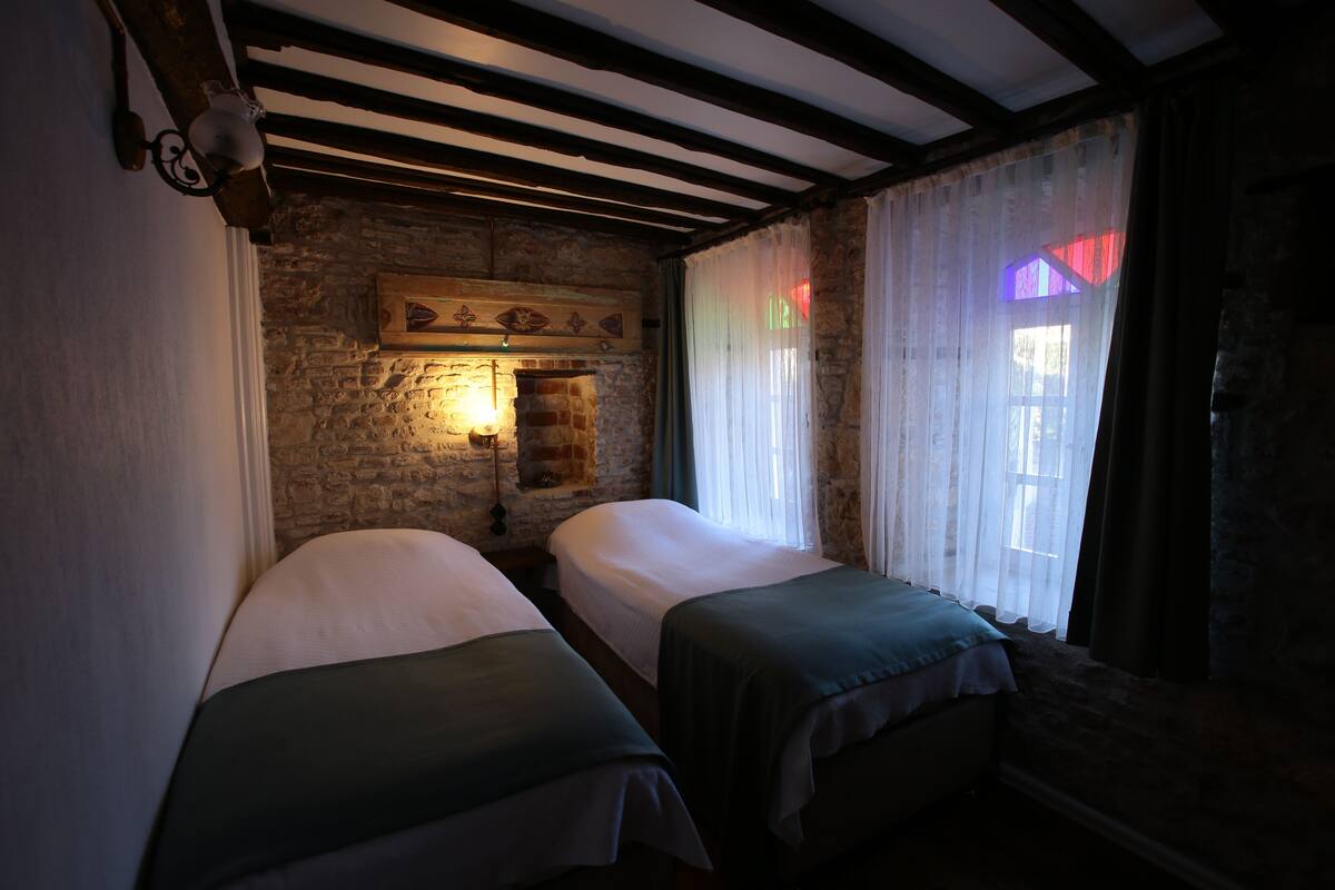 A room with two beds, a stone wall, a wooden ceiling, and a window with sheer curtains.