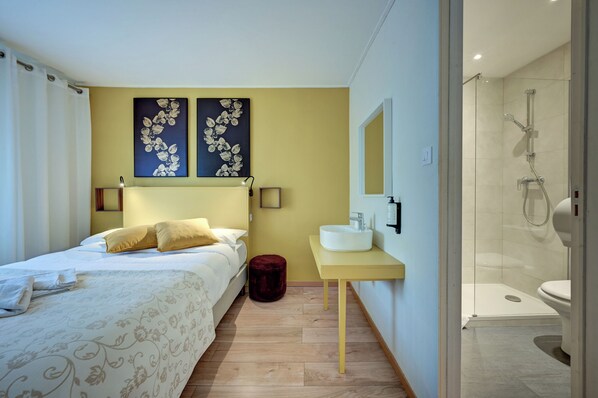 A hotel room with a bed, a bedside table, a small stool, a bathroom with a sink and toilet, and a shower. - Hotel Paul (Aix-en-Provence)