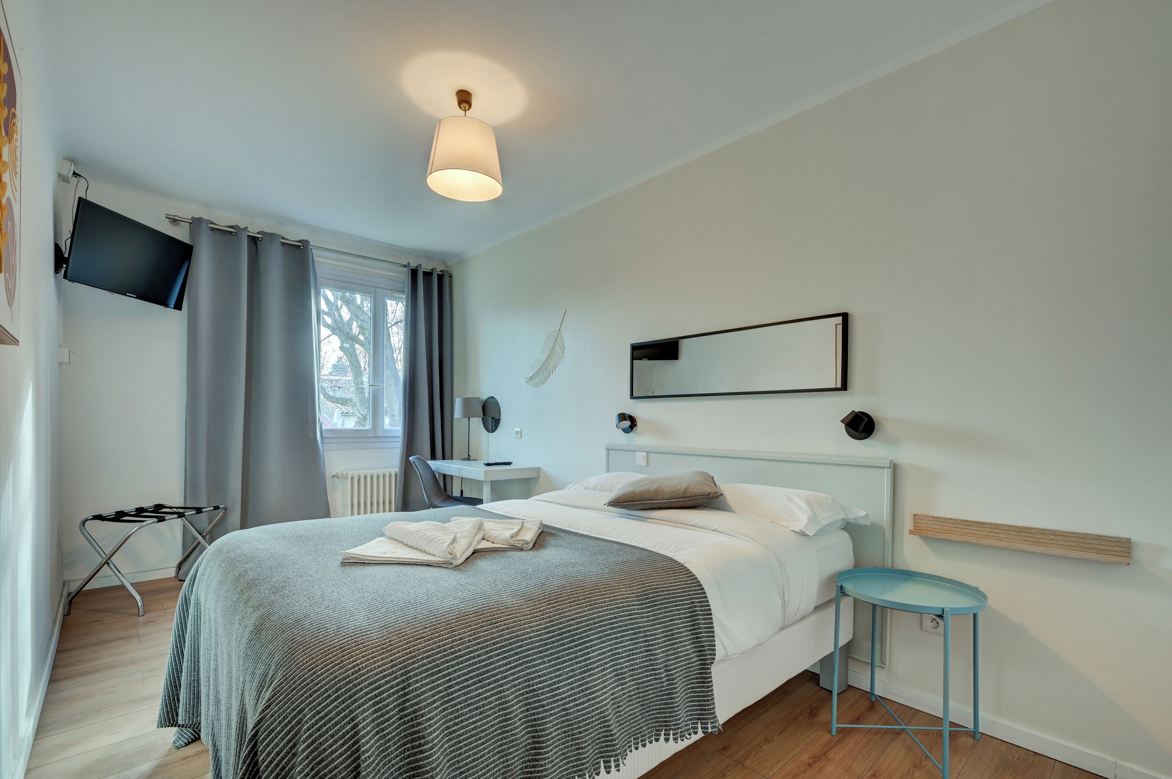 A modern hotel room with a bed, a desk, a chair, a TV, and a window with curtains.