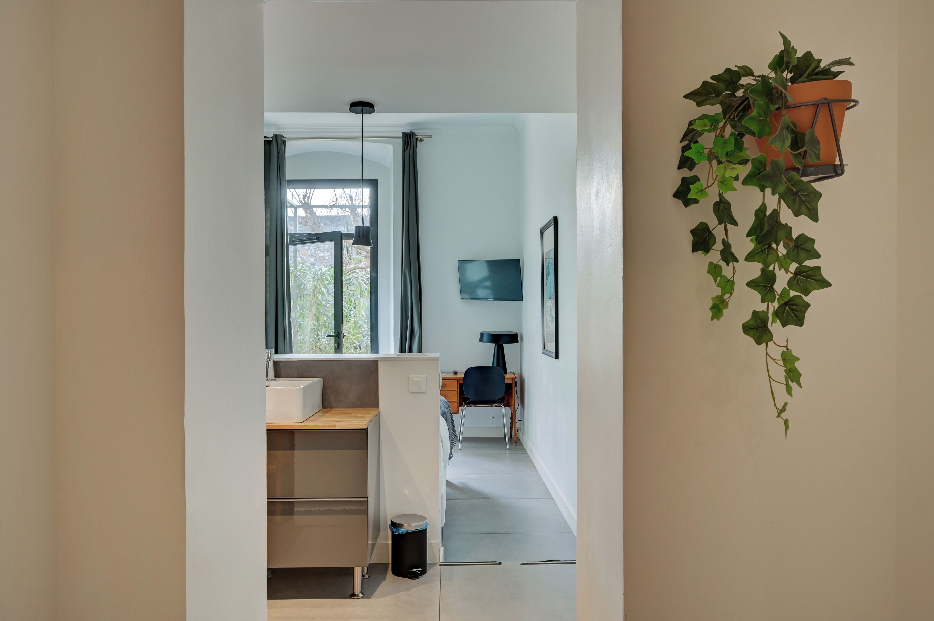 Une salle de bain moderne et minimaliste, équipée d’une douche, d’un lavabo et de toilettes. On y trouve une fenêtre avec des rideaux, une plante en pot et un tableau encadré accroché au mur.