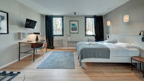 Un dormitorio moderno con cama, escritorio y silla, un televisor montado en la pared y dos ventanas con cortinas.
