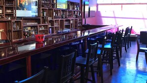 A bar area with wooden shelves stocked with bottles, a long bar counter, and several chairs. - Hotel Castillo (San José)