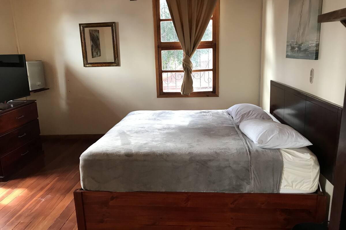 A neatly made bed with a wooden headboard, a dresser with a television, and a window with curtains.