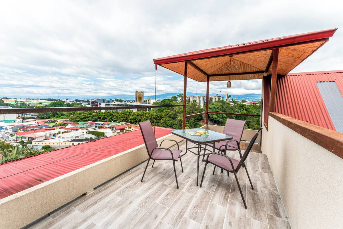 A balcony with a table and chairs, a red roof, and a view of a cityscape.
