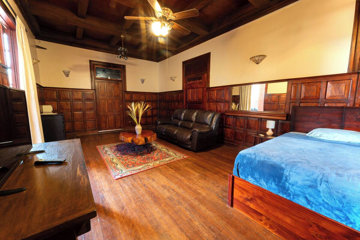 A room with a wooden ceiling, a bed with a blue bedspread, a leather sofa, a wooden table, and a rug.