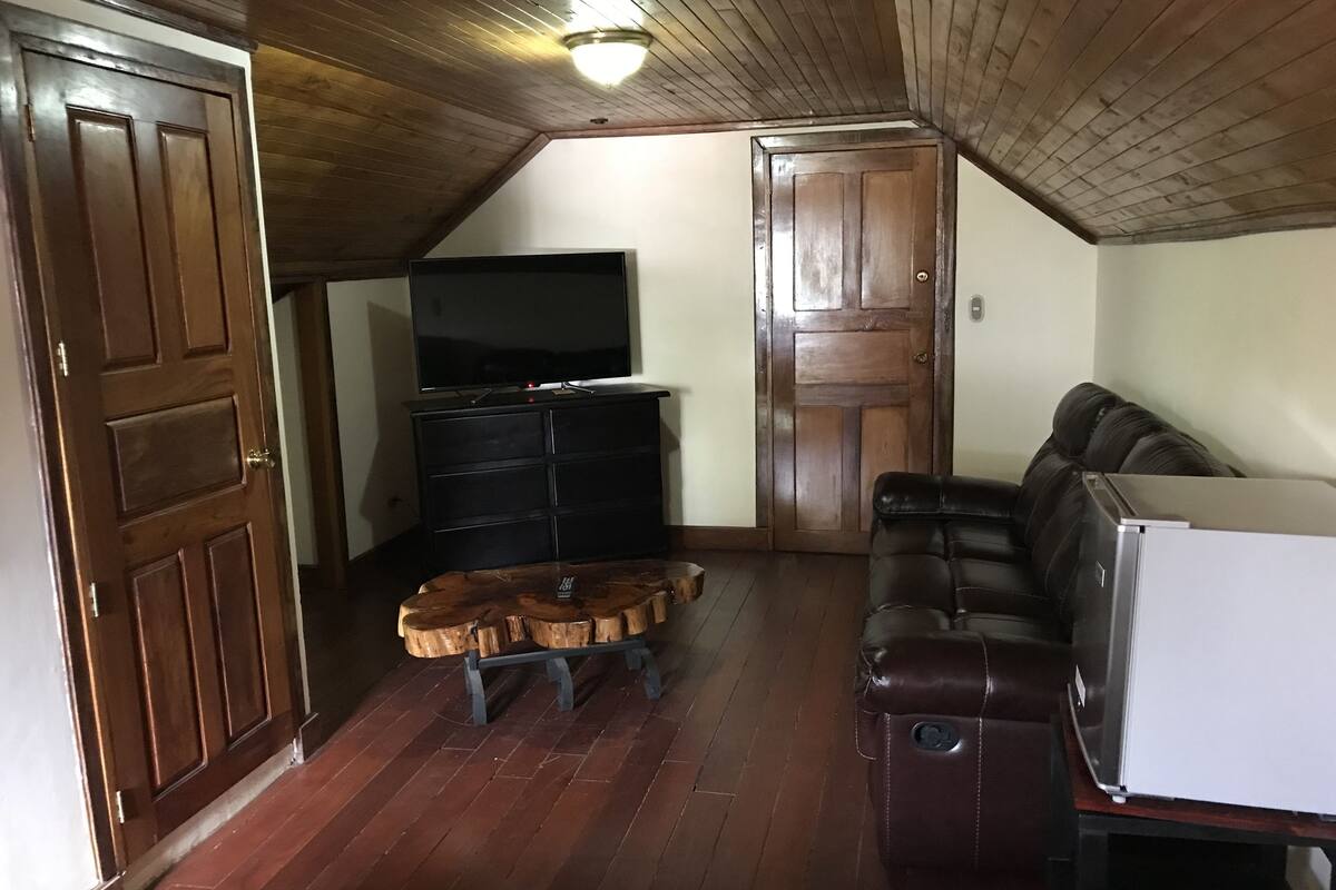 A room with a wooden ceiling, a television on a black stand, a leather sofa, a wooden coffee table, and a white refrigerator.