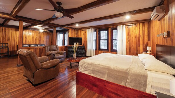 A room with a bed, two recliners, a wooden floor, a ceiling fan, and a TV. - Hotel Castillo (San José)