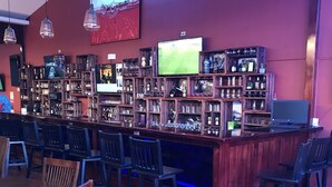 A bar area with wooden shelves stocked with bottles, a television mounted on the wall, and a seating area with chairs and a pink sofa. - Hotel Castillo (San José)