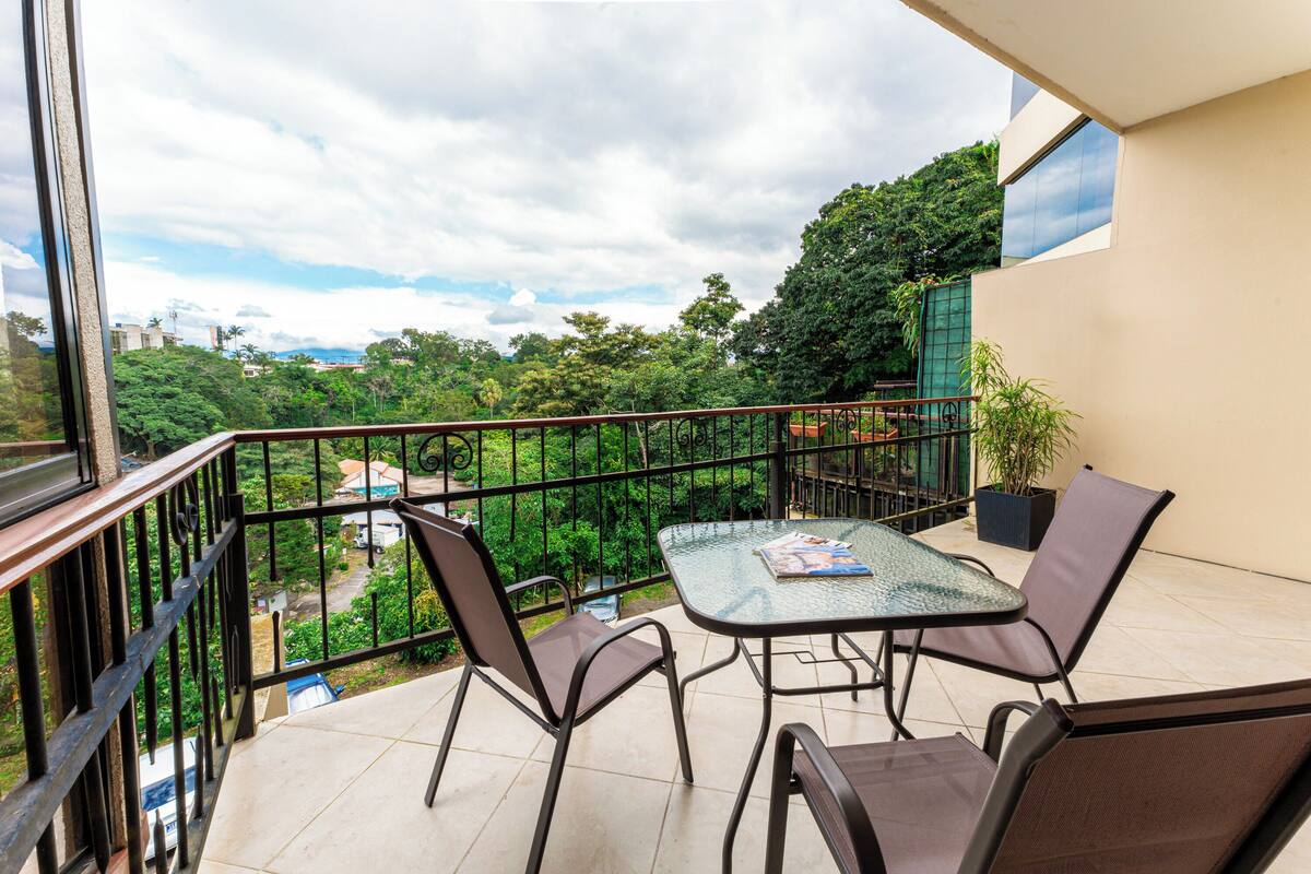 A balcony with a glass table and chairs, a potted plant, and a view of greenery and buildings.