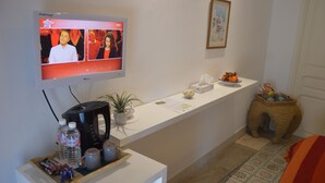 A hotel reception area with a flat-screen TV displaying a program, a coffee maker, water bottles, a plant, a small table with a bowl and tissues, and a decorative basket. - La Chambre Verte (Sidi Bou Said)