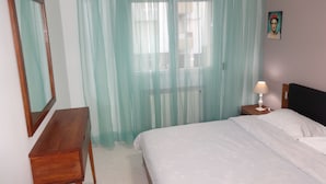 A bedroom with a bed, a bedside table with a lamp, a mirror, and sheer curtains.