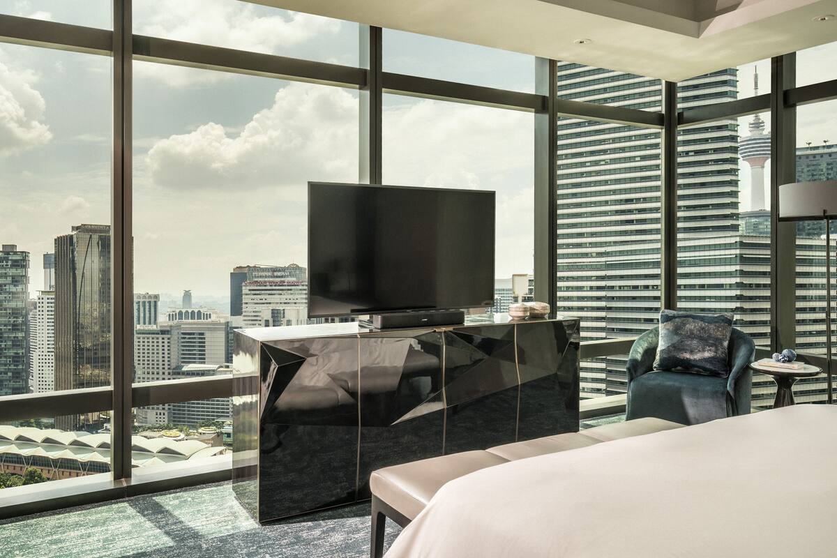 Une chambre d’hôtel moderne avec une grande télévision posée sur une table en verre, un lit confortable et une vue sur la ville grâce à de grandes fenêtres.