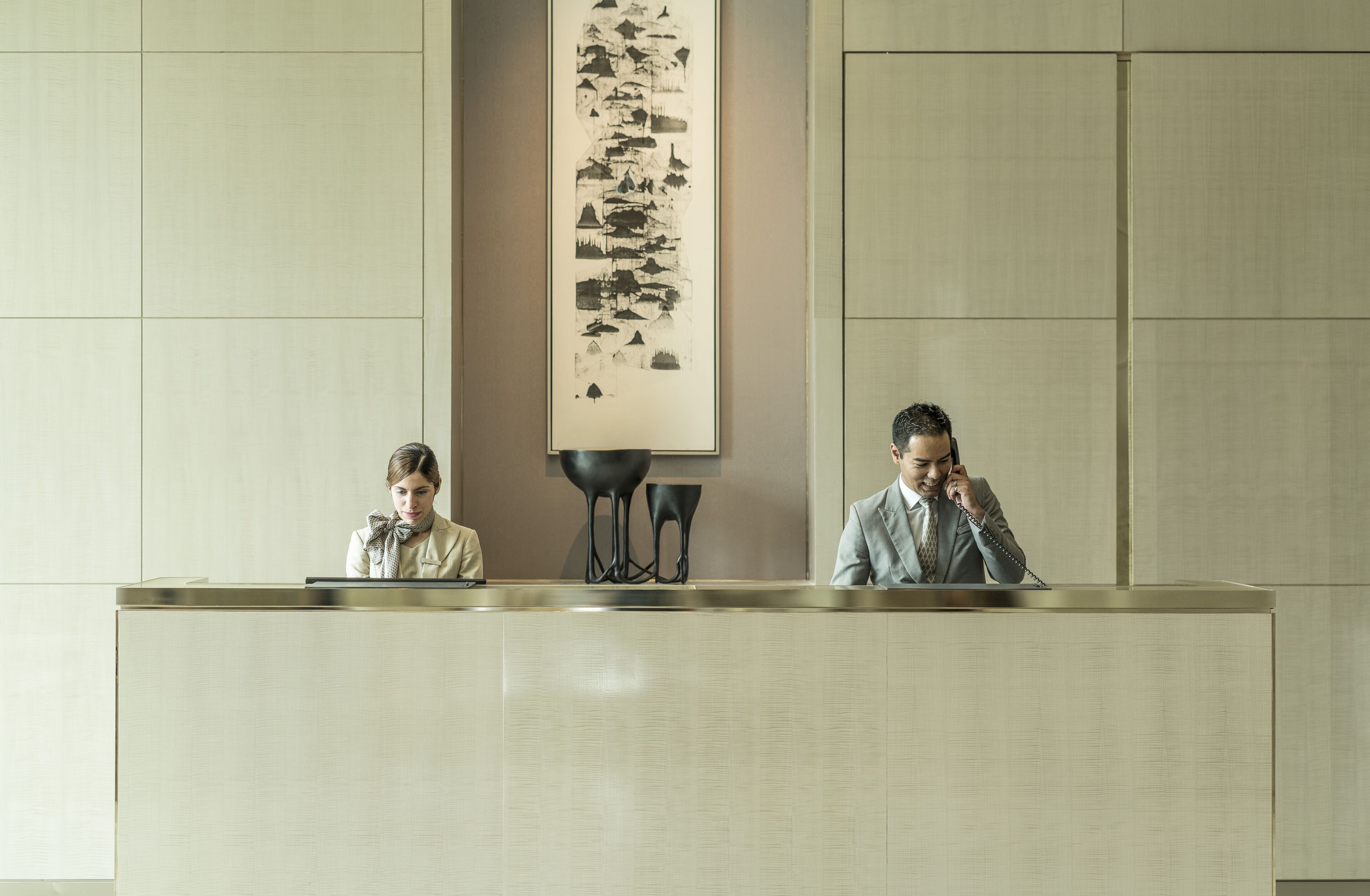 Un mostrador de recepción con dos personas sentadas, una obra de arte abstracta vertical sobre ellos y un diseño interior moderno y minimalista.
