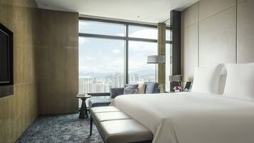 A modern hotel room with a large bed, a seating area with a small table, and a view of the cityscape through large windows.