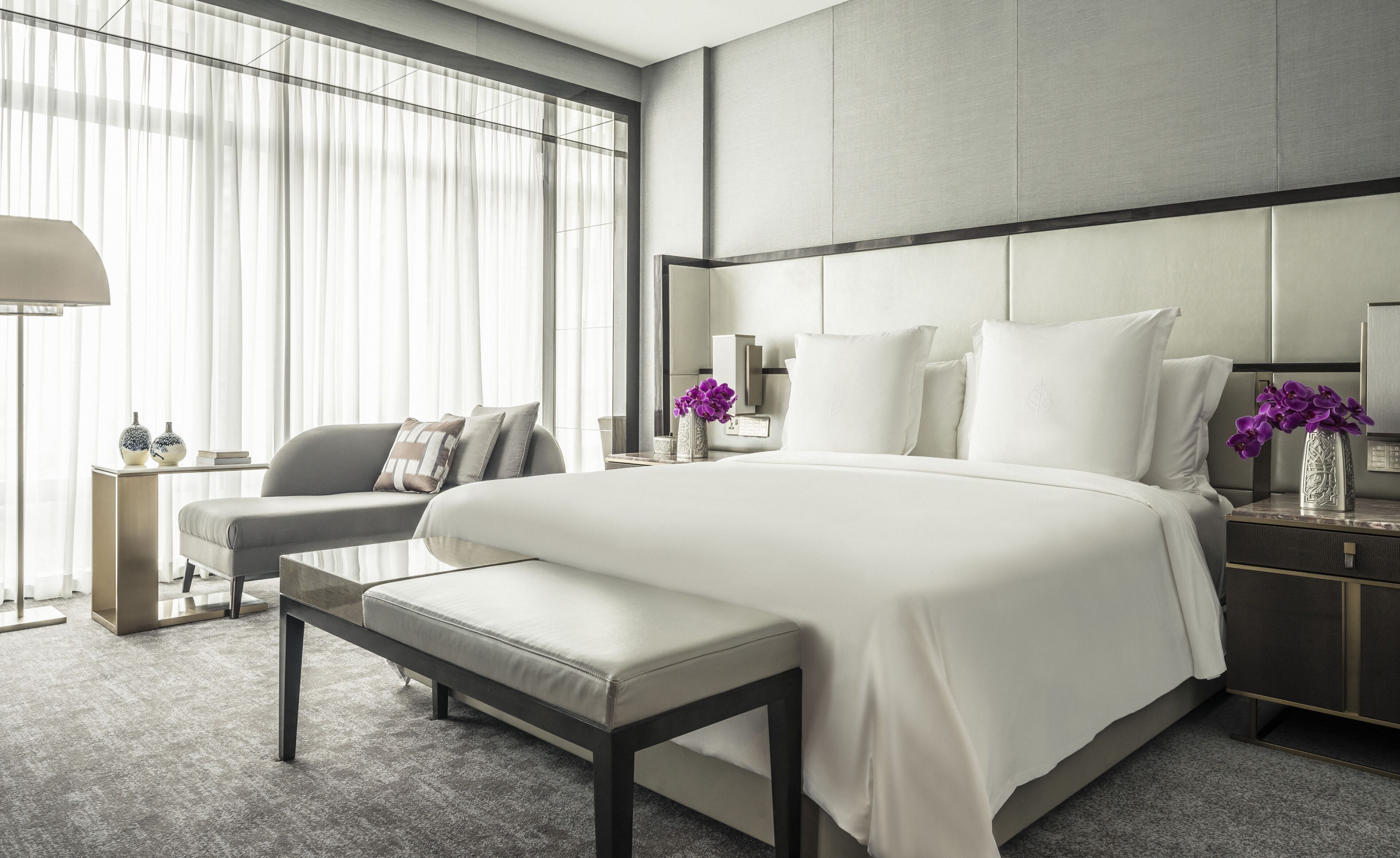 A modern hotel room with a large bed, a seating area with a bench, and a nightstand with a vase of flowers.