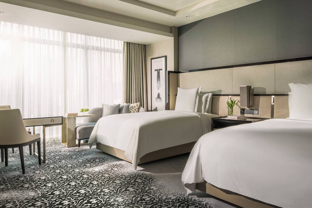 Une chambre d’hôtel avec deux lits, un bureau, une chaise, une grande fenêtre avec des rideaux, un tableau encadré au mur et une moquette à motifs.
