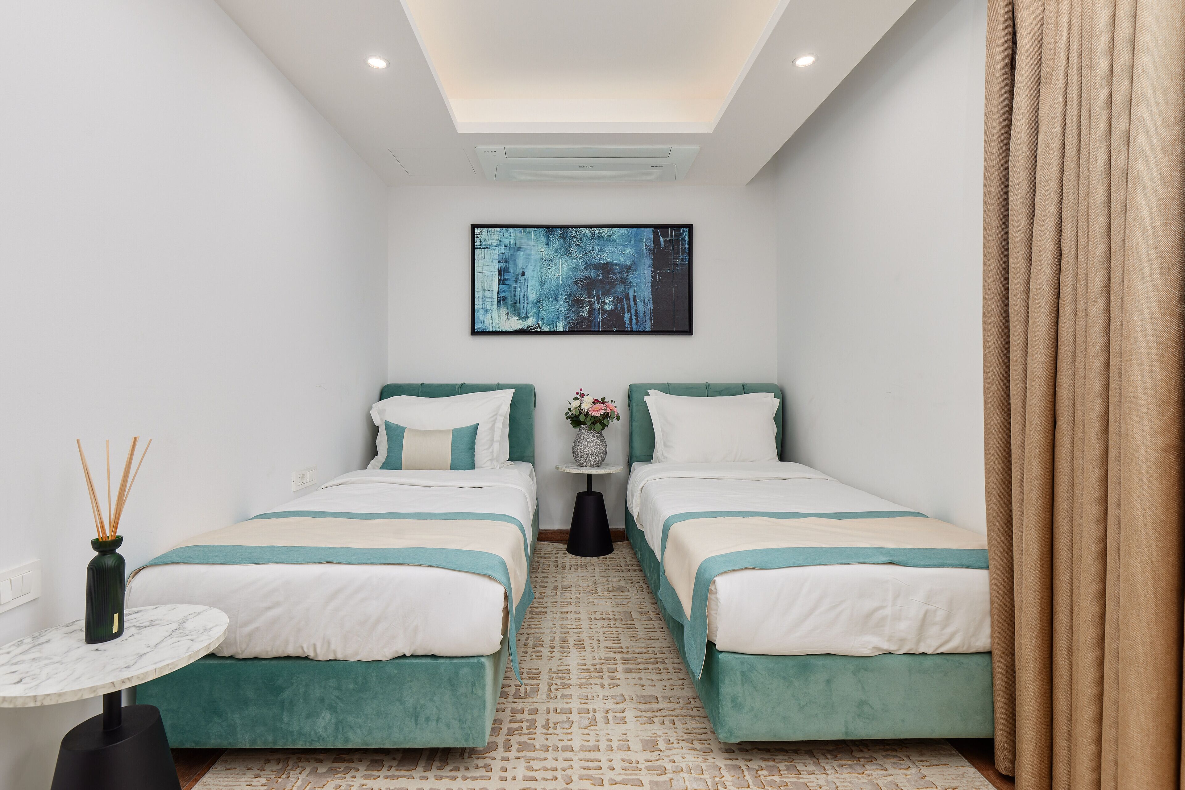A hotel room with two beds, a small table, a vase with flowers, and a painting on the wall.