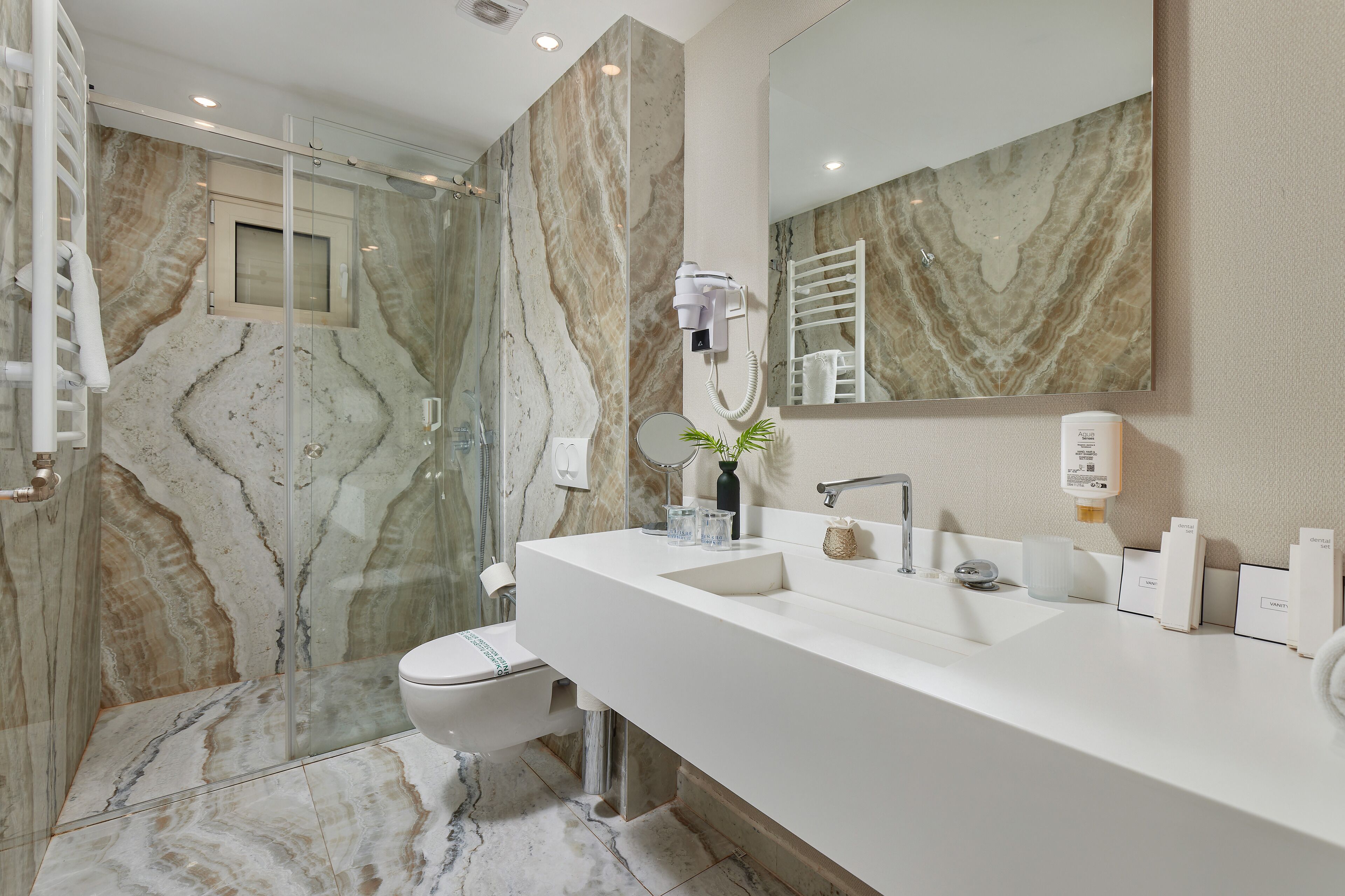 Un baño moderno con pared de ducha de mármol, un espejo grande, un lavabo blanco y un secador de pelo de mano fijado a la pared.