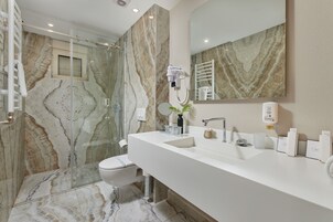 Un baño moderno con pared de ducha de mármol, un espejo grande, un lavabo blanco y un secador de pelo de mano fijado a la pared.