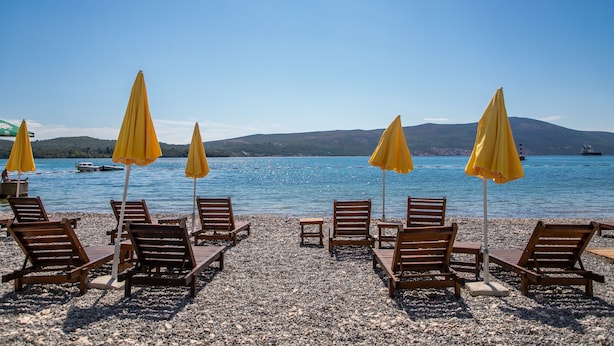 Una playa con sillones de madera y sombrillas amarillas, una playa de guijarros y un mar tranquilo con barcos a lo lejos.