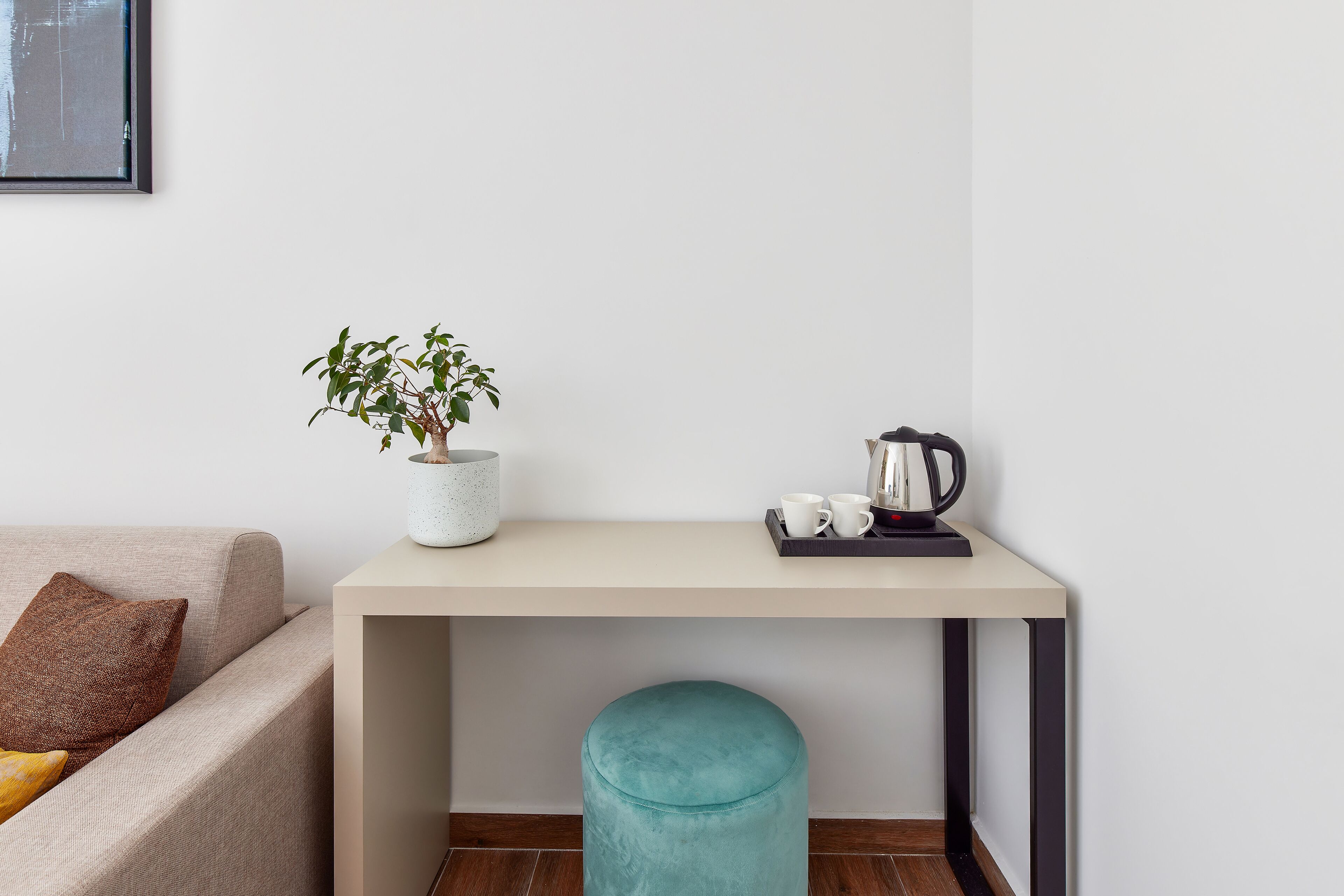 A room with a beige sofa, a small round table with a plant, two cups, and a kettle.