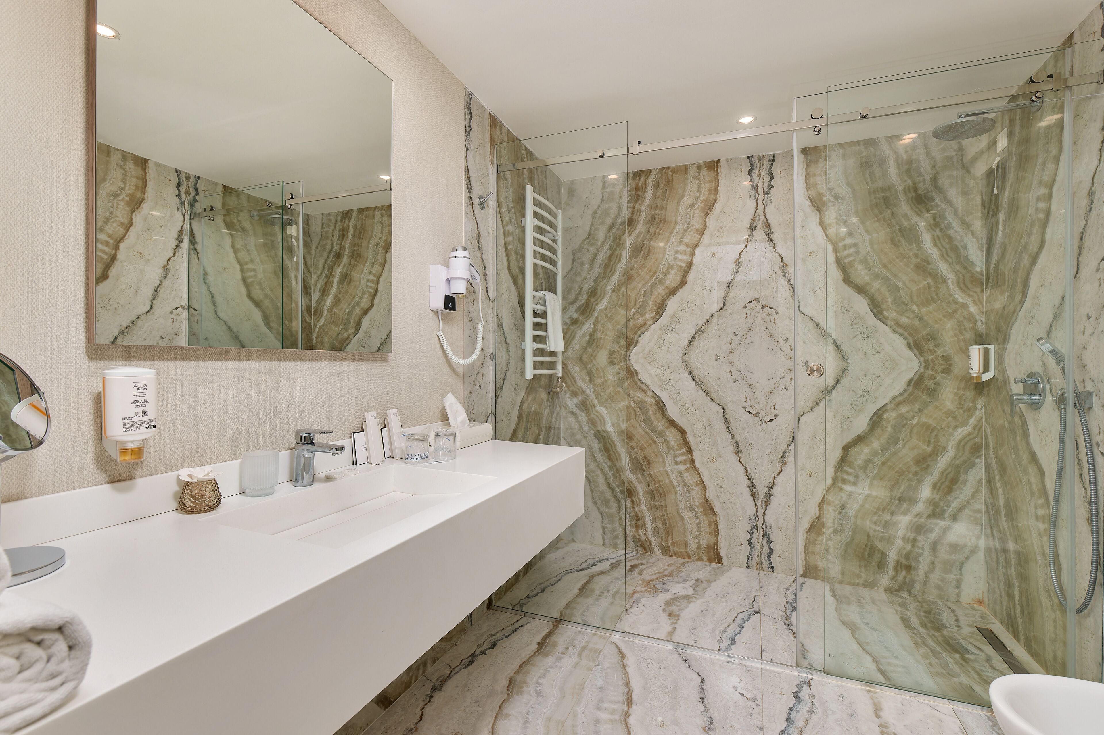 Un baño moderno con pared de ducha de mármol, ducha con cerramiento de vidrio, lavabo blanco con espejo y un perchero para toallas.