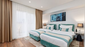 Habitación de hotel con dos camas, un ventanal con cortinas translúcidas, un cuadro en la pared y una mesita con lámpara.