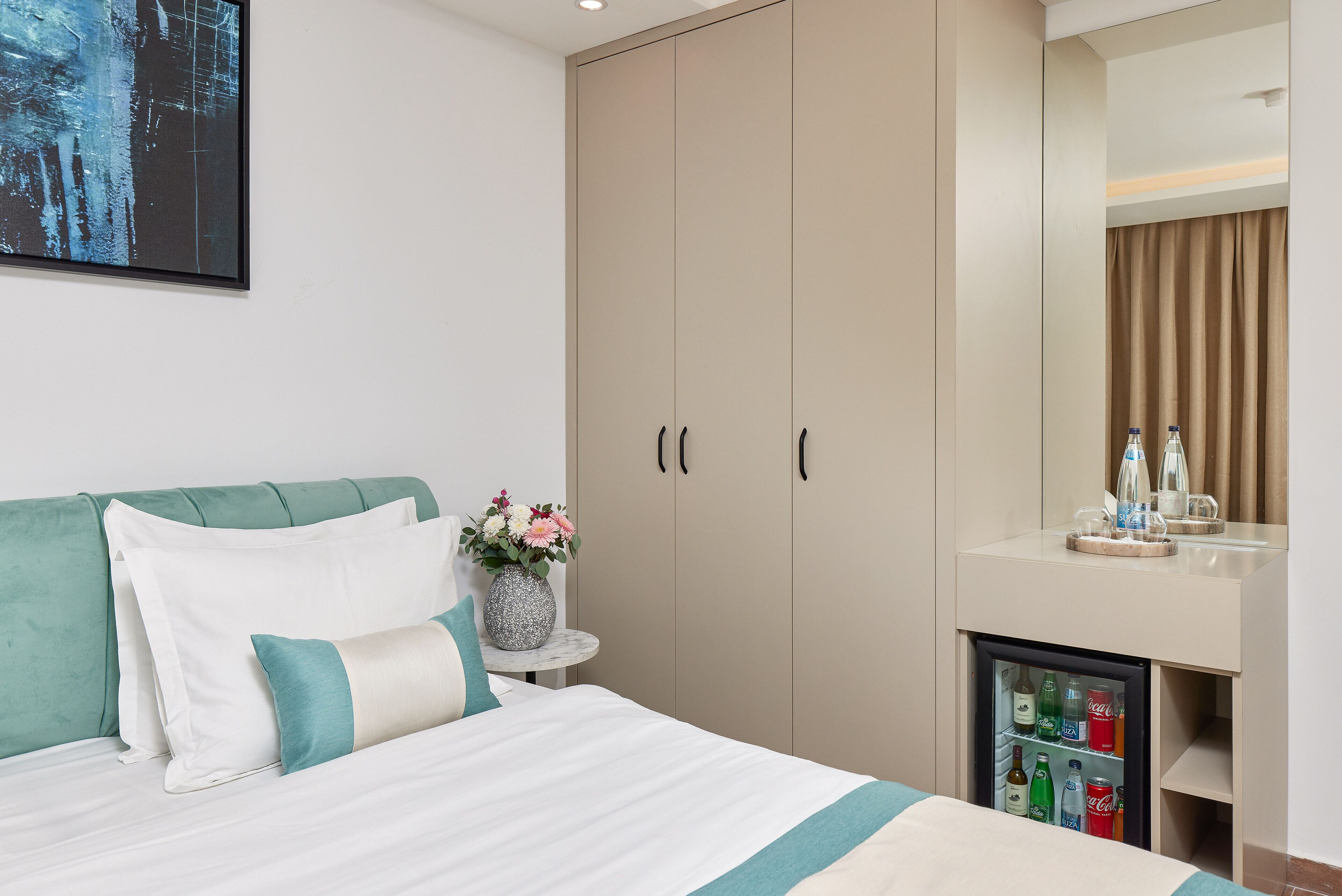 A hotel room with a bed, a small table with a vase of flowers, a minibar, and a large wardrobe.
