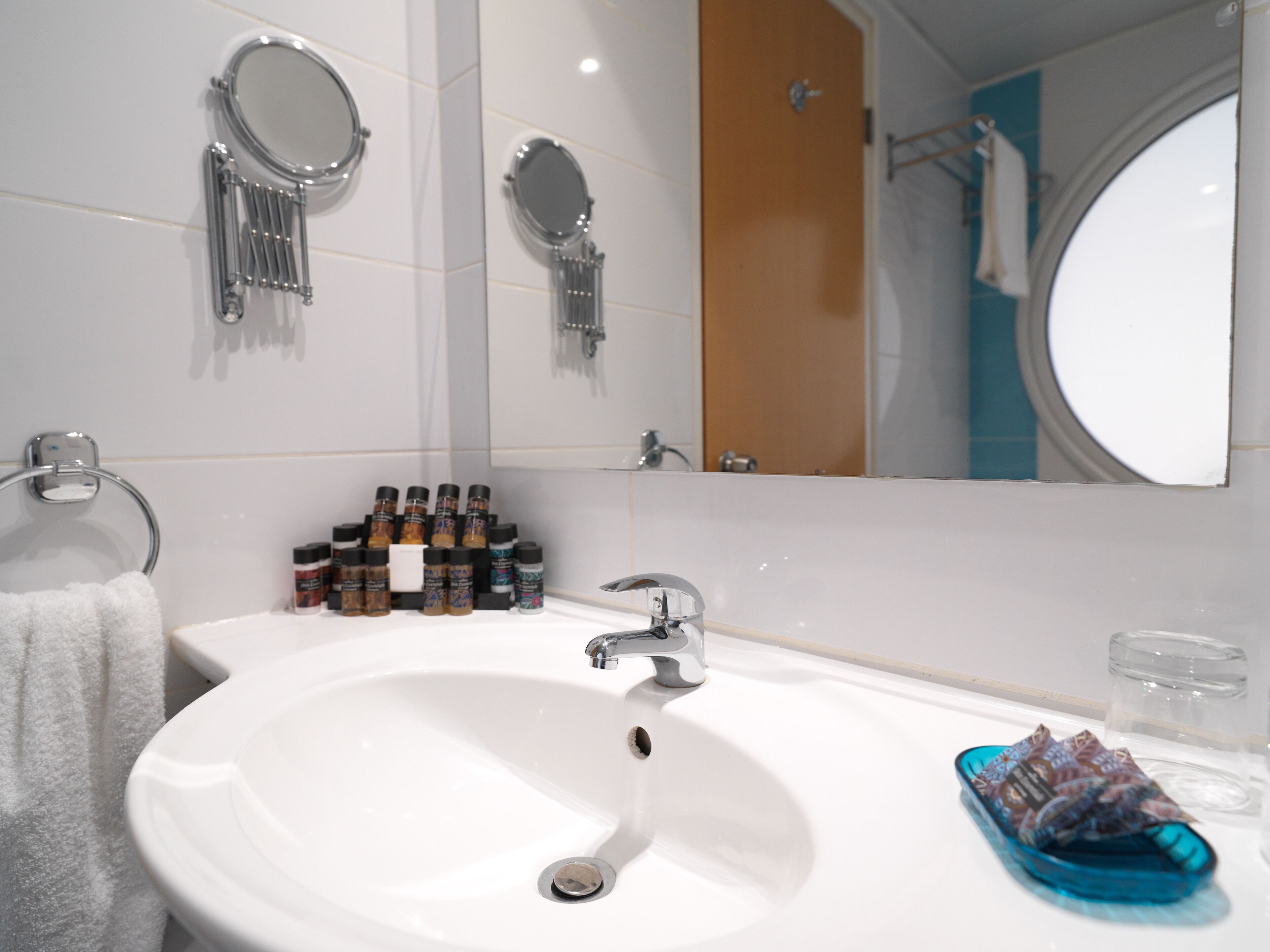 A bathroom with a white sink, two round mirrors, a towel rack, and a shelf with various toiletries.