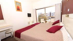 A hotel room with a bed, a desk, a chair, and a view of the city through large windows. - Hotel Kings Annex (Paphos)