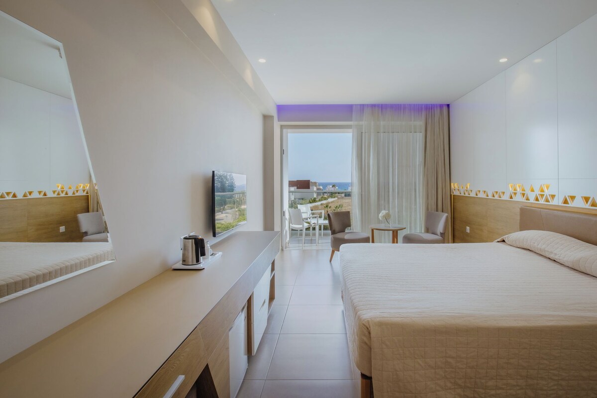 A hotel room with a bed, a desk with a coffee maker, a TV, and a view of the beach.