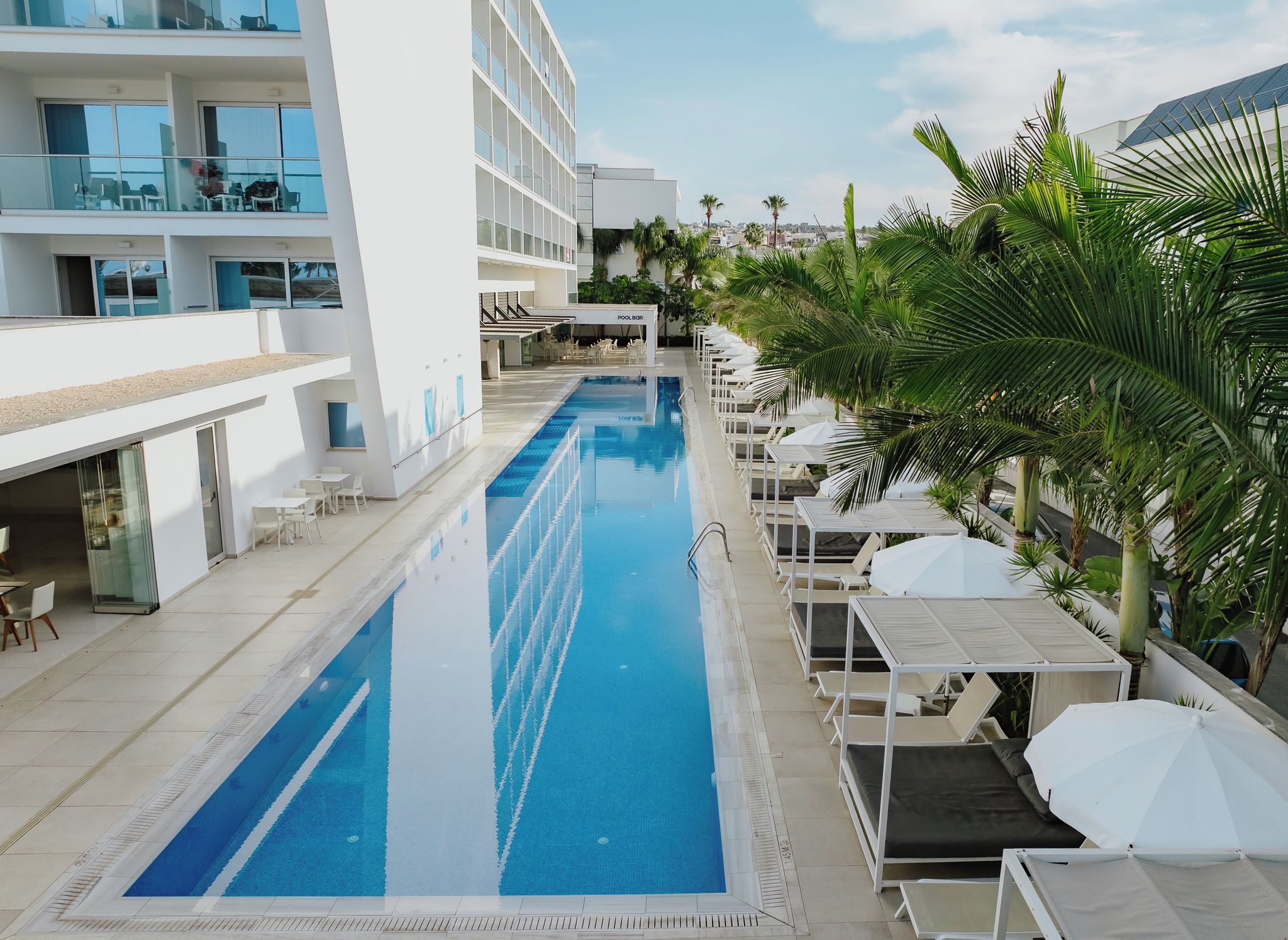 A swimming pool with lounge chairs and umbrellas, surrounded by palm trees and a modern building with multiple floors and balconies.
