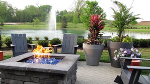 A stone fire pit with a fire, surrounded by Adirondack chairs and potted plants, set against a backdrop of a lake and fountain. - Renaissance Columbus Westerville-Polaris Hotel (Westerville)