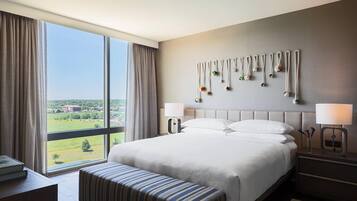 Habitación de hotel con una cama grande, un banco, una mesita de noche y vistas al exterior.