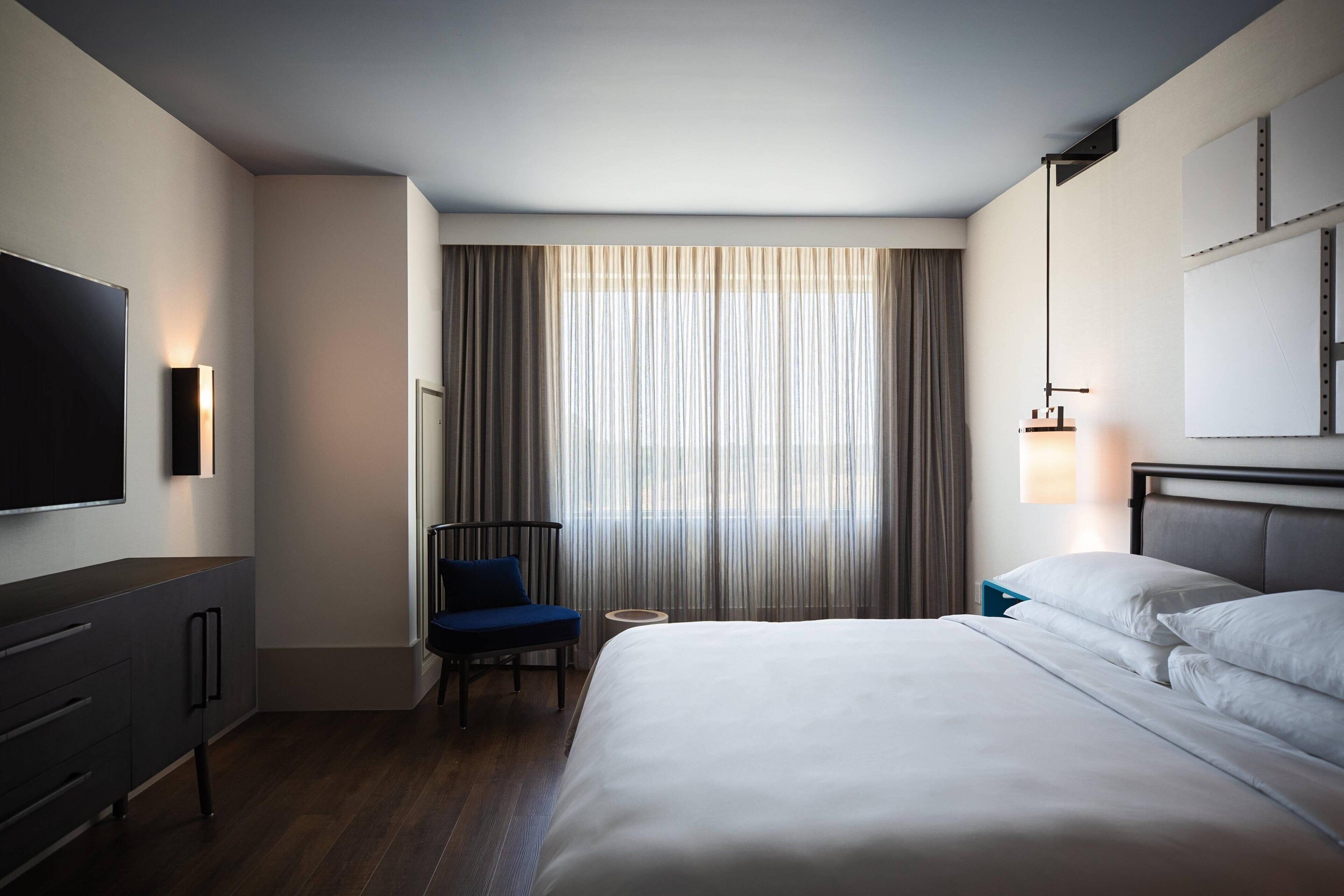 A hotel room with a bed, a TV, a chair, a small table, and a window with curtains.