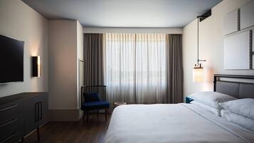 Habitación de hotel con cama, televisión, una silla, una mesita y una ventana con cortinas.