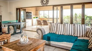 A living room with a sofa, coffee table, and a pool table. There is a balcony with a dining area and a kitchen in the background. - Seaview Luxury (Jeffreys Bay)