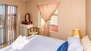 A hotel room with a bed, a chair, a nightstand with a lamp, a window with curtains, and a view of the outdoors. - Tuscany On Sea (Great Brak River)