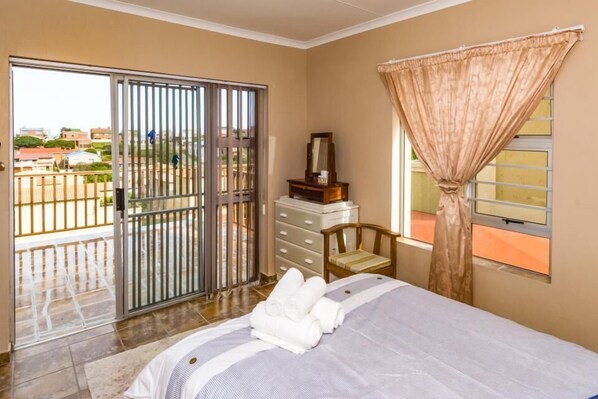 Une chambre d’hôtel comprenant un lit, une commode, une chaise, une fenêtre avec des rideaux et une porte-fenêtre coulissante donnant sur un balcon.