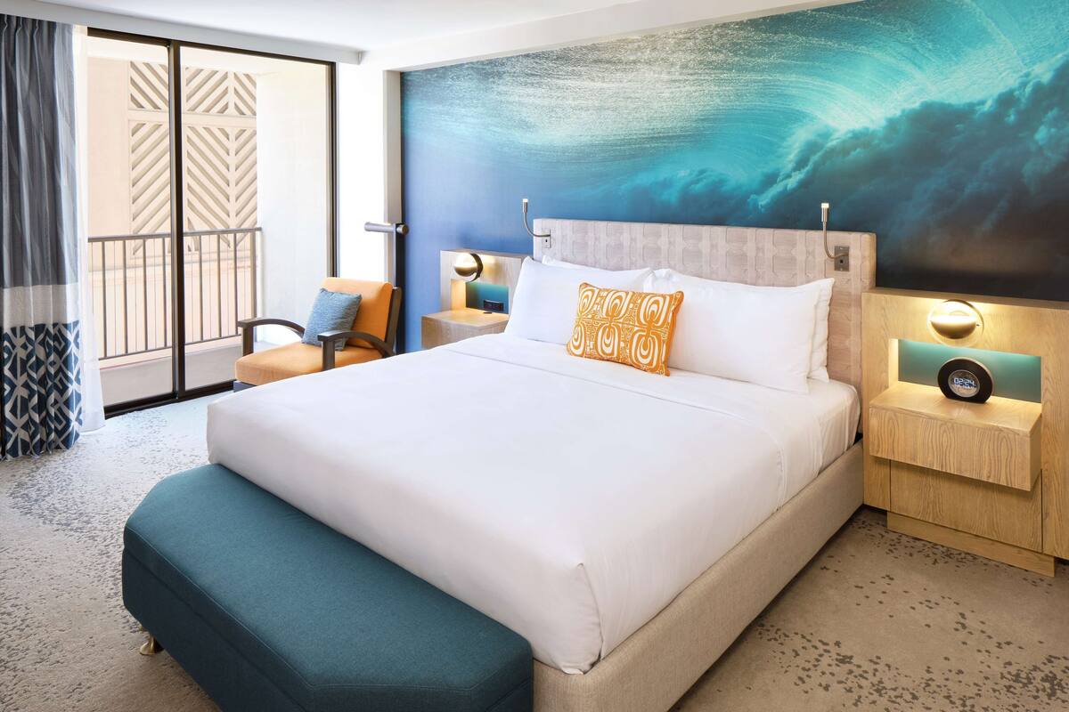 A hotel room with a large bed, a nightstand with a clock, a chair, and a view of the ocean through a sliding door.