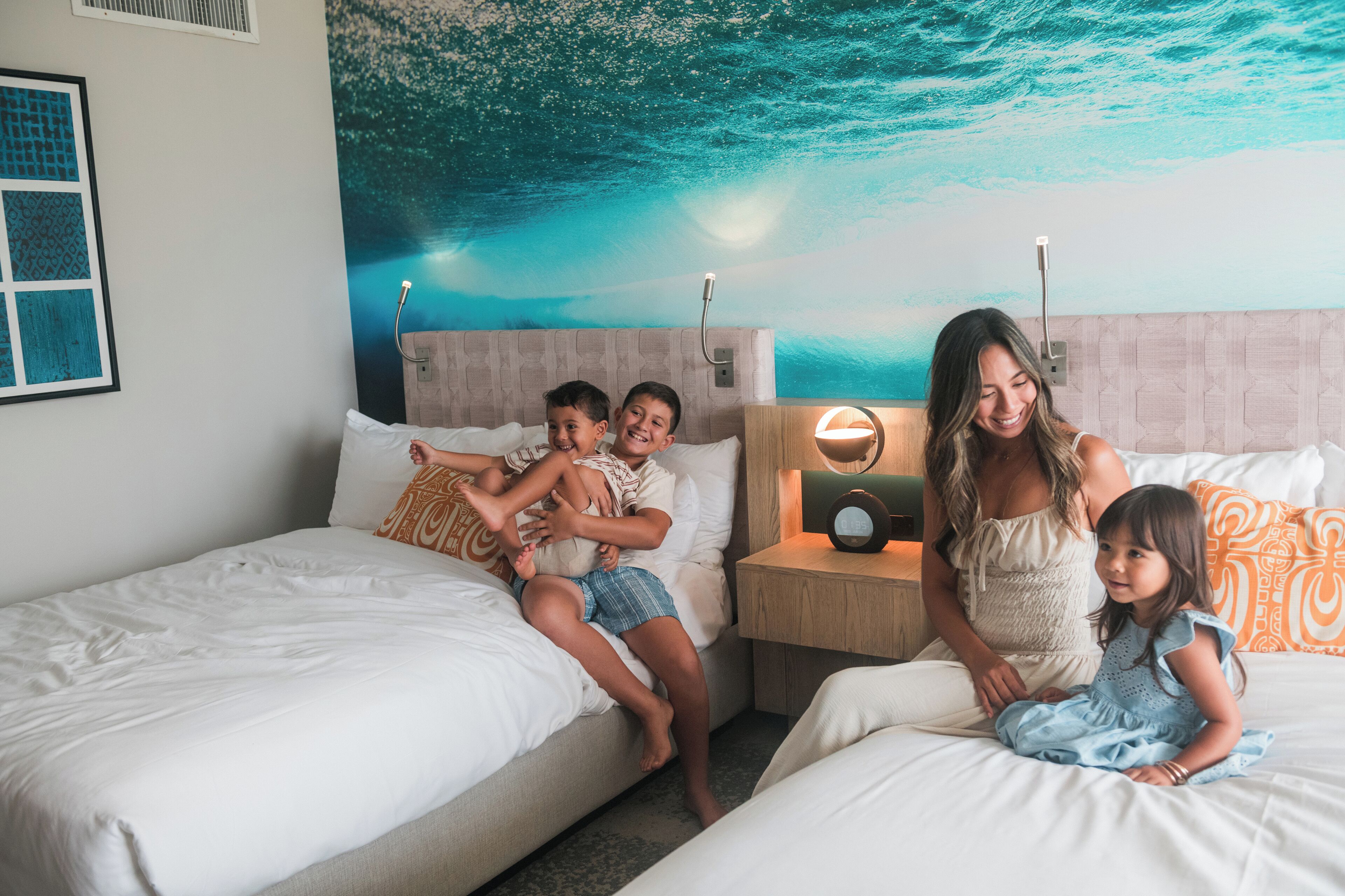 A hotel room with two beds, a mural of the ocean on the wall, and a bedside table with a lamp.