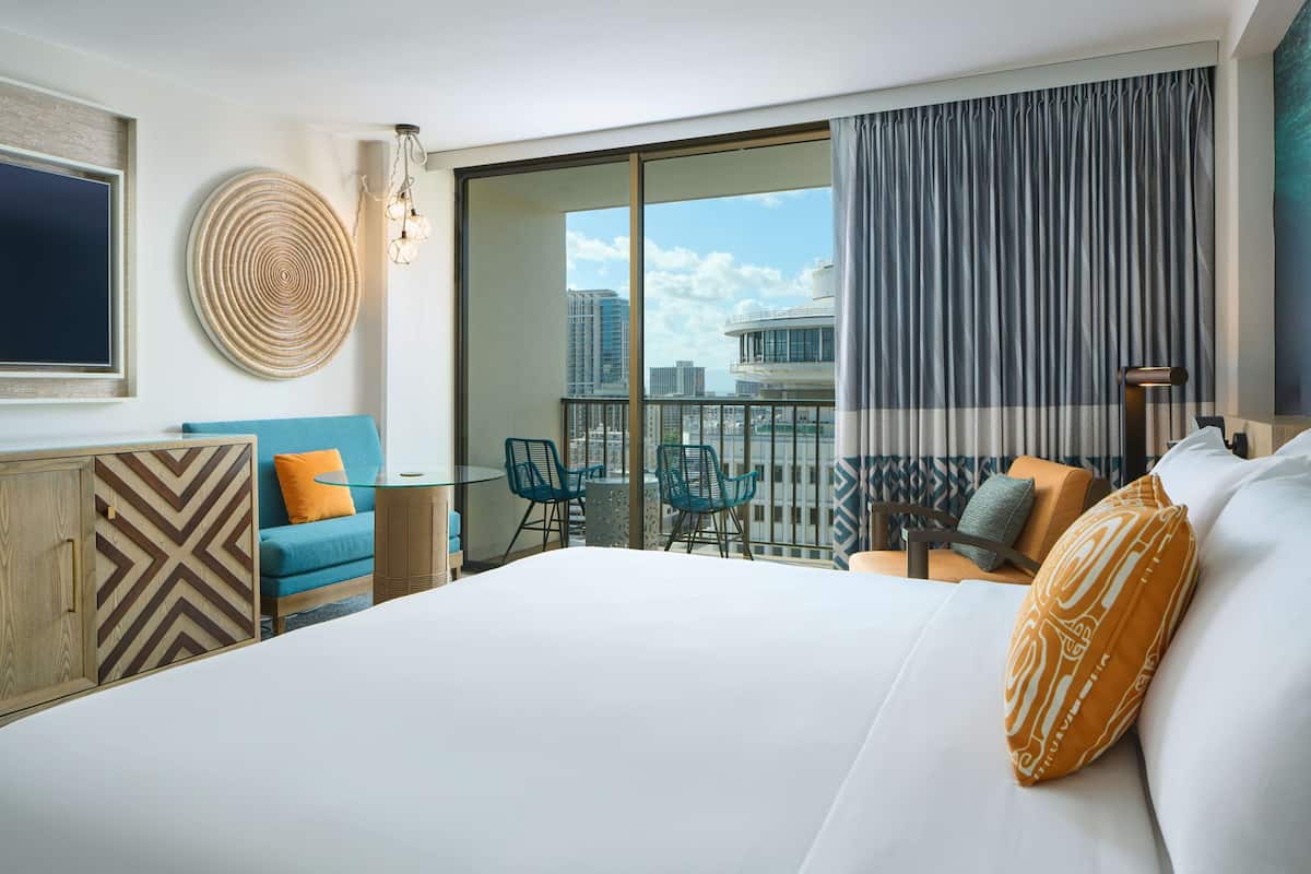A modern hotel room with a large bed, a balcony with a view of the city, a round woven wall art piece, and a blue sofa with orange cushions.