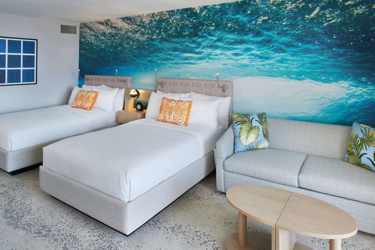 A hotel room with two beds, a sofa, a coffee table, and a large ocean-themed wall mural.