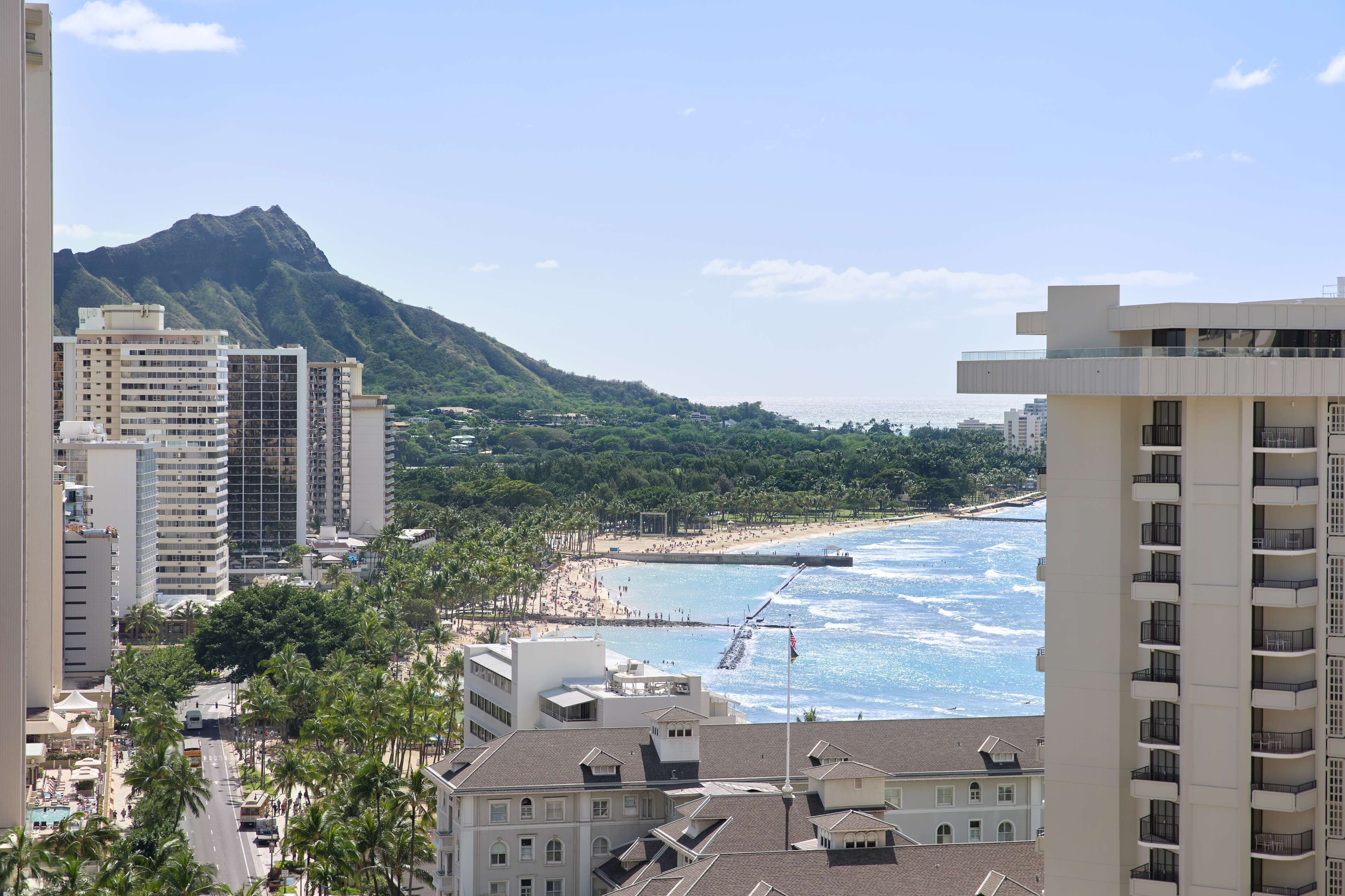 Paisagem costeira com edifícios altos, uma praia e uma montanha ao fundo.