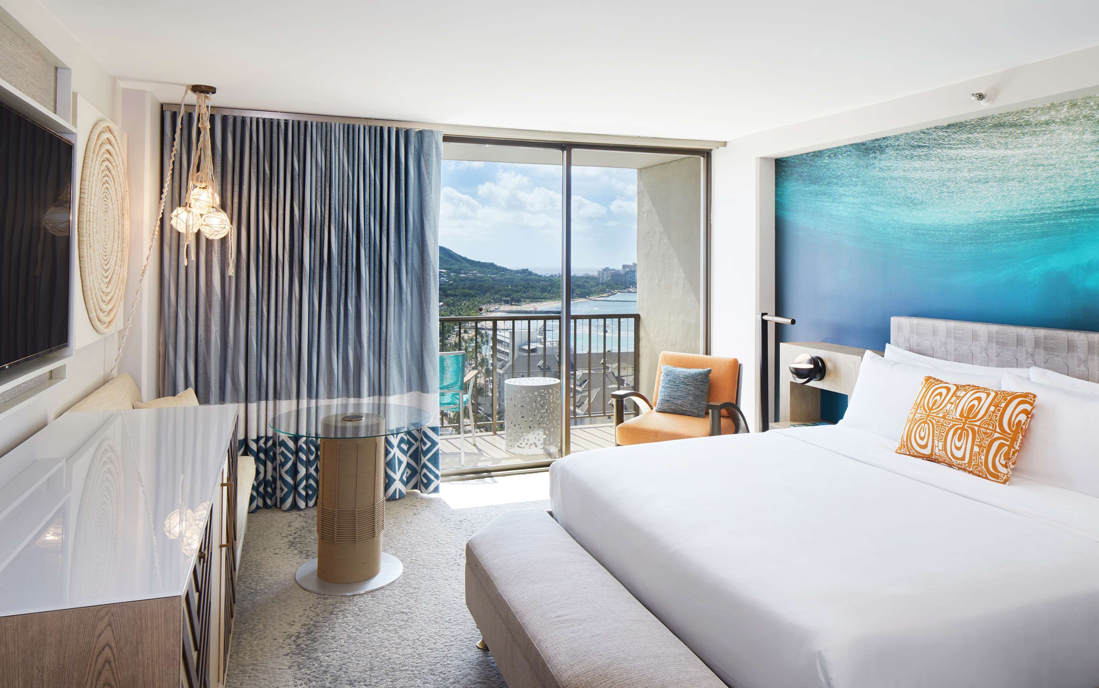 A modern hotel room with a large bed, a TV, a balcony with a view of the sea, and a glass table.