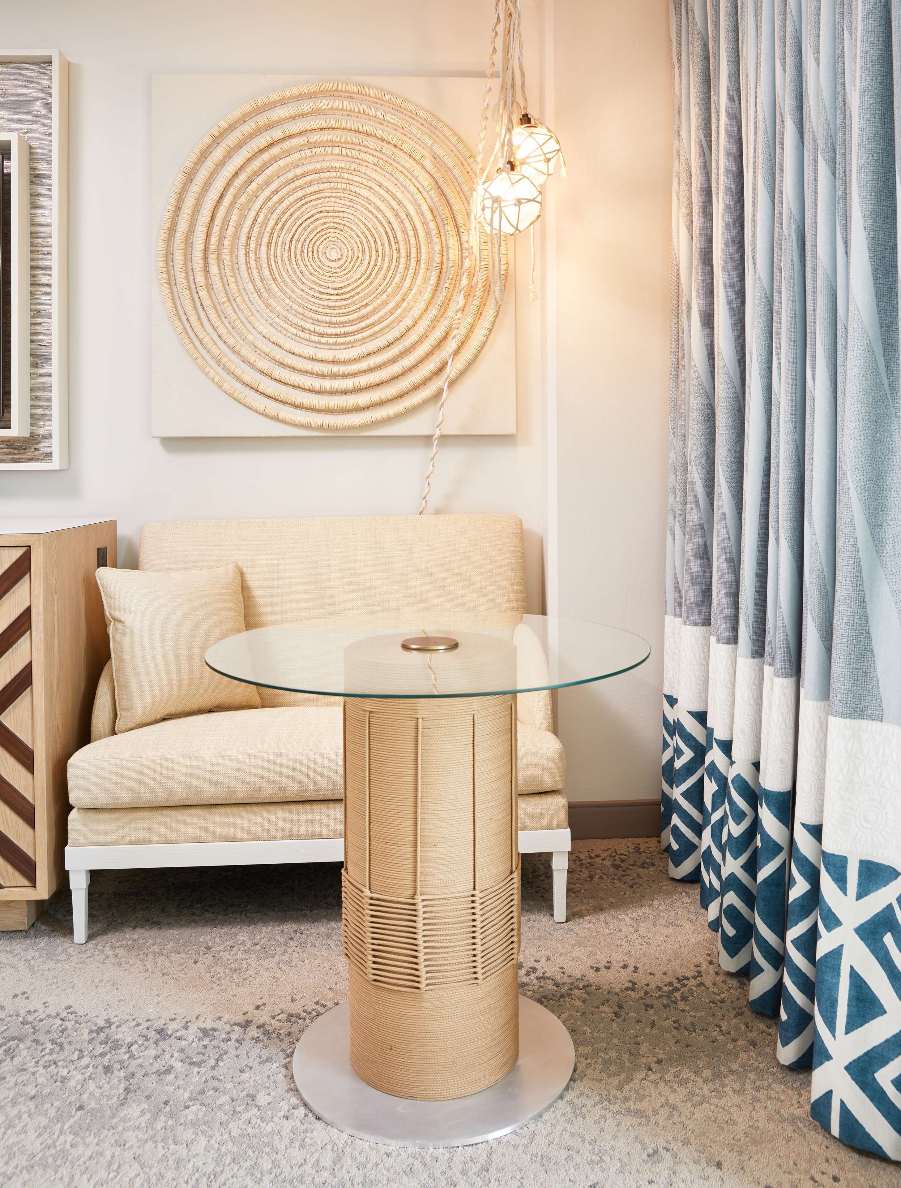 A modern living room with a beige sofa, a glass-top coffee table, a woven side table, a wall-mounted woven artwork, and patterned curtains.