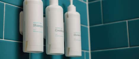 Three white bottles of coconut lime shampoo, conditioner, and shower gel on a teal tiled wall.
