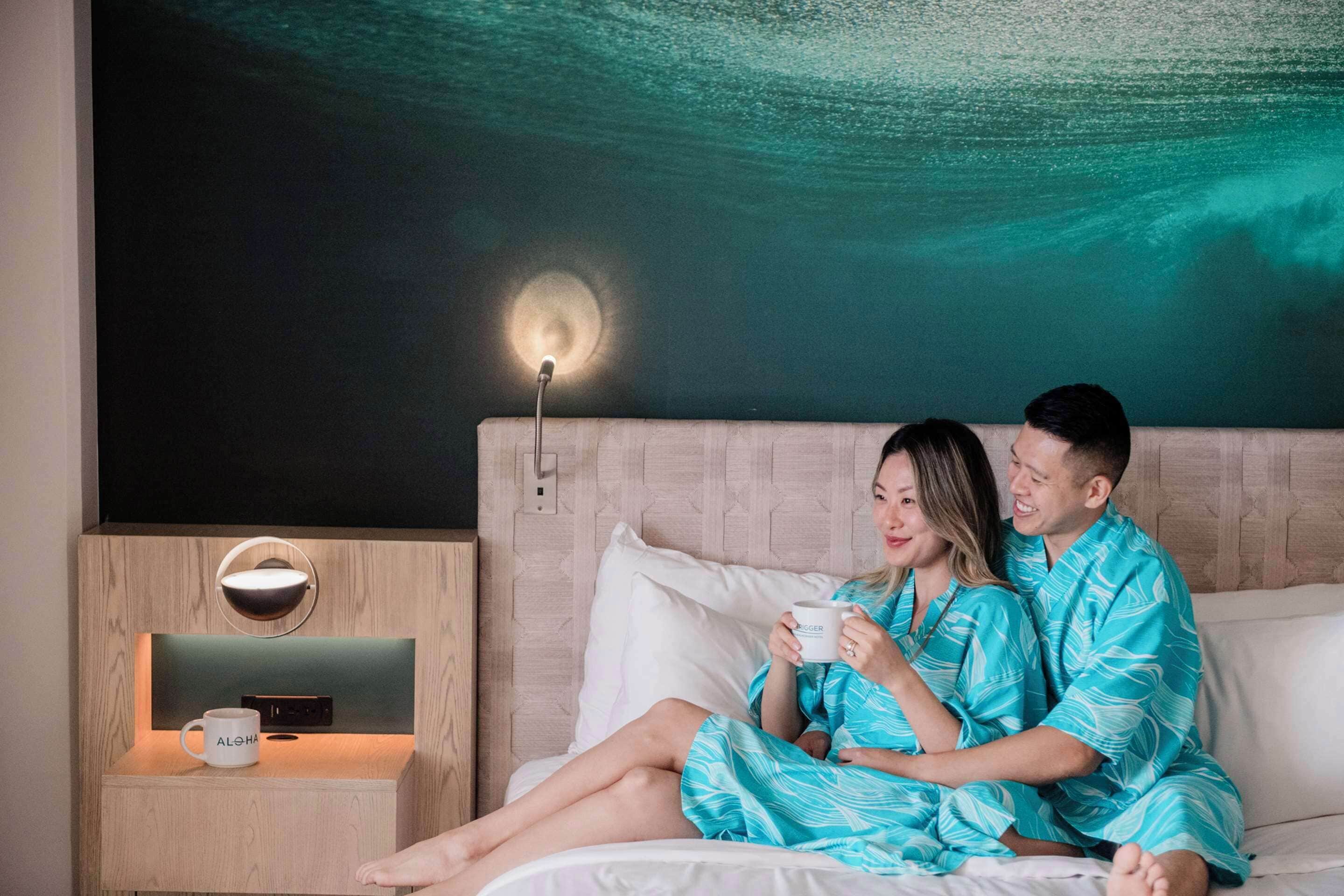 Un couple, vêtu de pyjamas bleus assortis, est assis sur un lit. Une table de chevet se trouve à côté du lit, sur laquelle sont posées des tasses.
