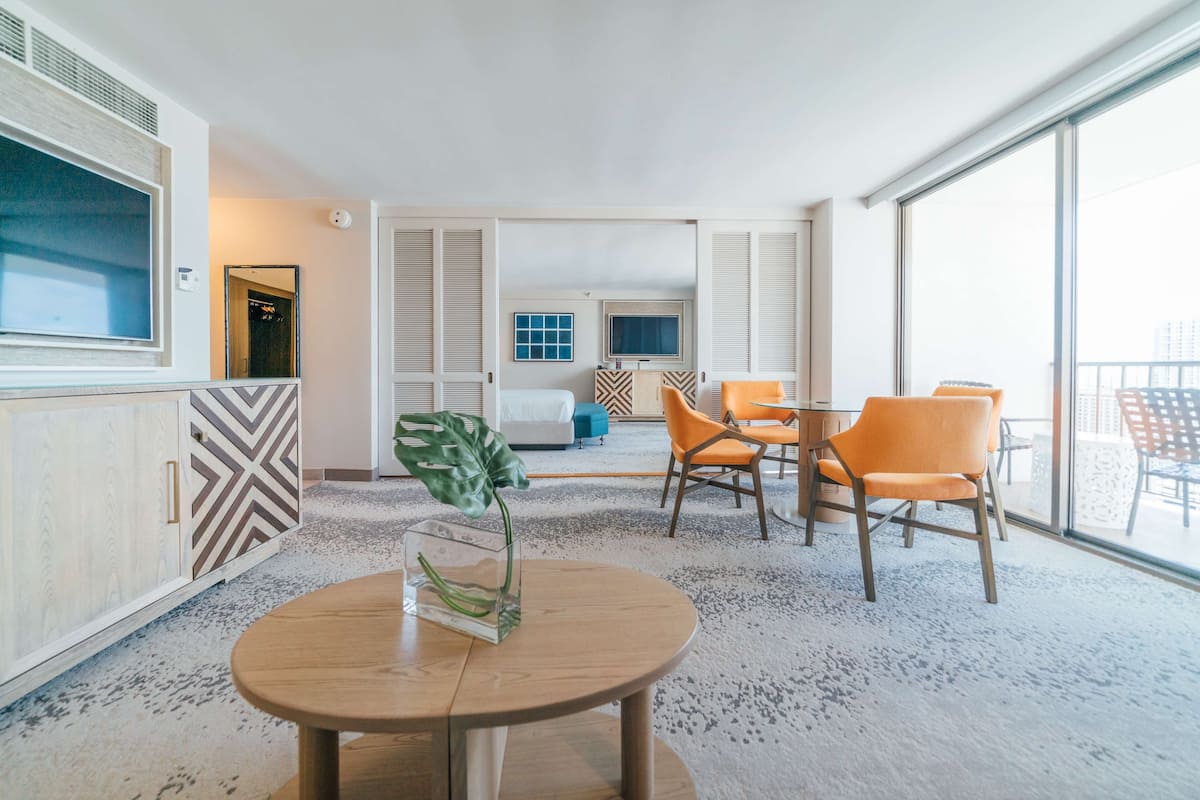 A modern hotel room with a large window, a round wooden table with a vase, two orange chairs, a bed, and a TV.
